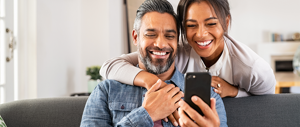 Couple using cell phone, man is sitting woman is hugging him